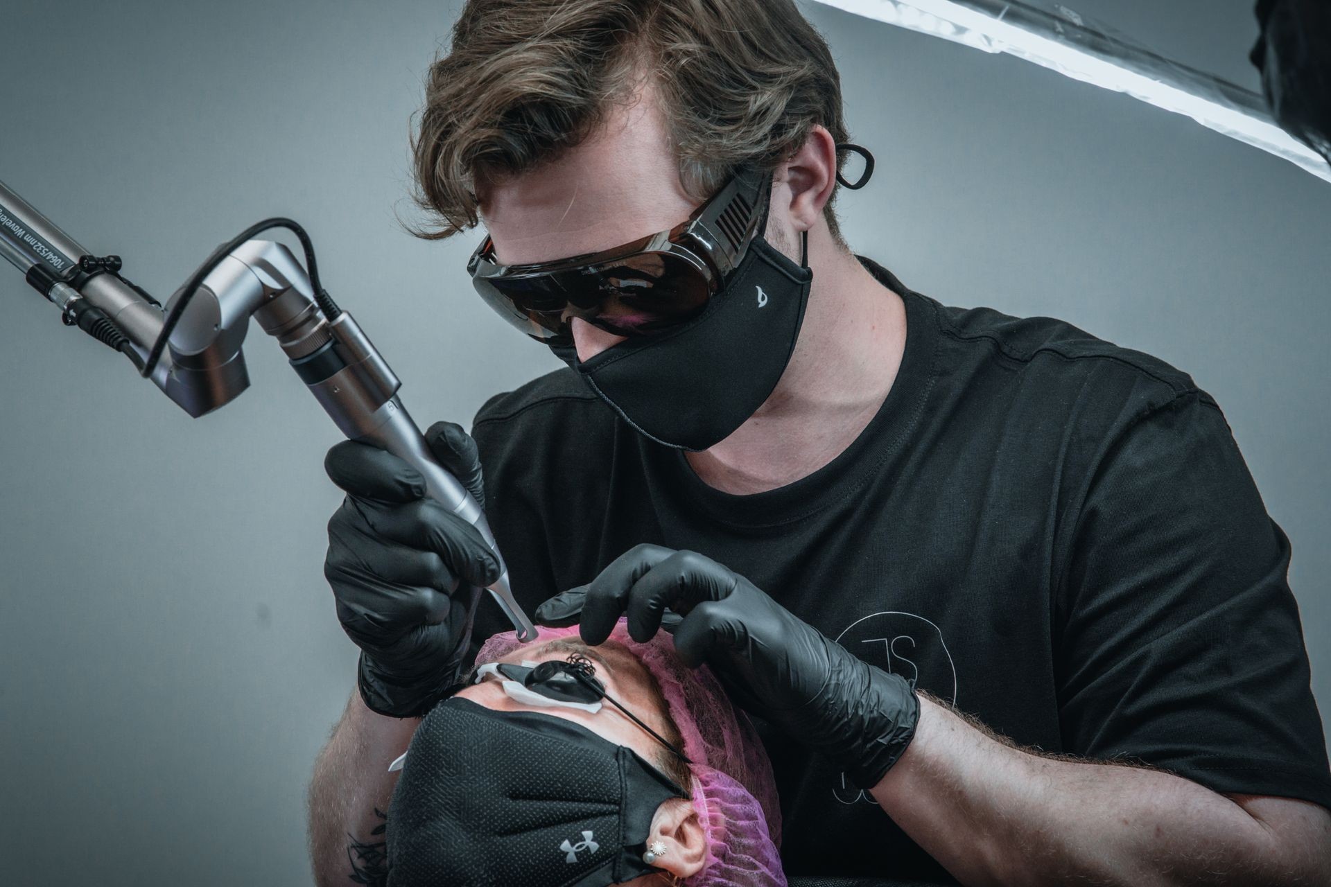 Tattoo artist with gloves and mask using a tattoo machine on a client's arm in a studio.